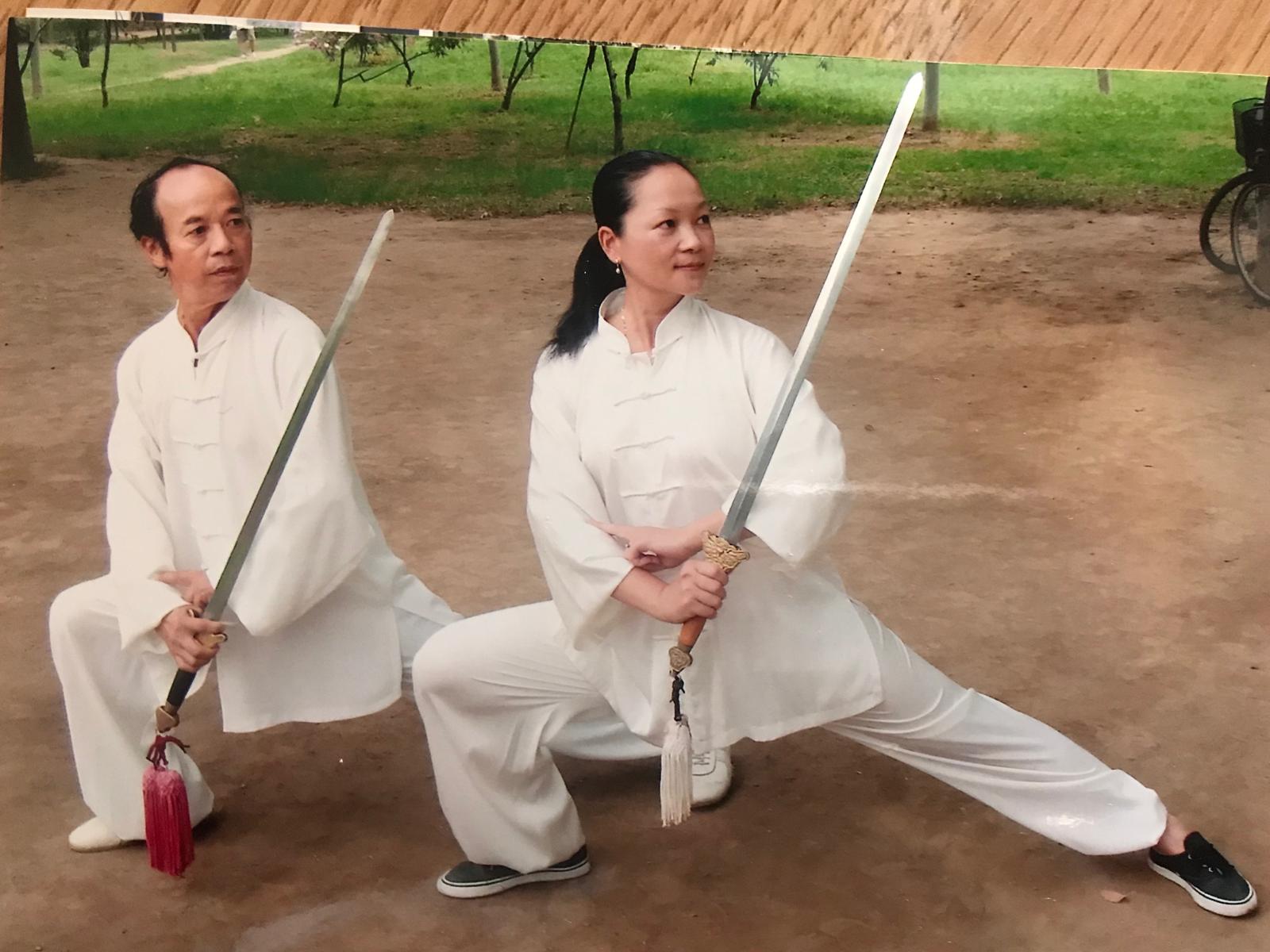 Two Tai Chi practitioners in white training clothes holding swords in a deep, balanced lunge